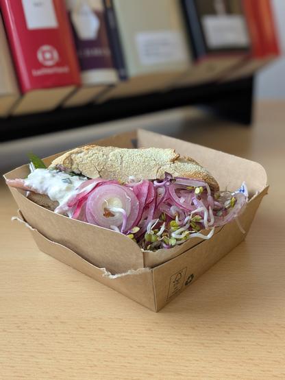 Ein Fischbrötchen mit Matjes, Remoulade, roten Zwiebeln und Sprossen auf einem Schreibtisch im Gerichtshaus Lübeck. Im Hintergrund ein Handapparat mit Kommentarliteratur und Formularhandbüchern.