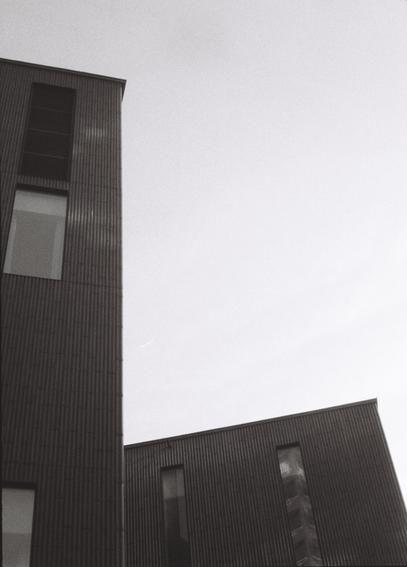Black-and-white photograph of two dark, boxy buildings forming a kind of L shape against the bright sky. The buildings have simple façades with a rifled texture and plain, rectangular windows matching the shapes of the buildings.