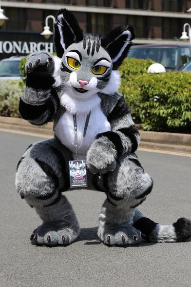 Full-body photo of a tabby cat fursuiter. She is looking directly into the camera, crouching, and reaching up with one paw in a maneki-neko pose. She is outside a con hotel in a parking lot on a sunny day.