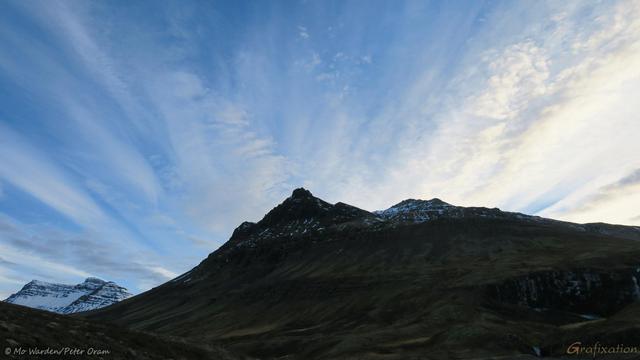 A colour photo of a snow-capped mountain range beneath a blue sky streaked with lines of cloud. A pointed peak is in the centre, and the stripes of hazy cloud seem to radiate from it.