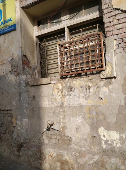 A photo of a window with a small window AC attached to it and barricaded with rusty iron grating. The wall surrounding the window is dilapidated and is flaking cement, exposing the bricks underneath.