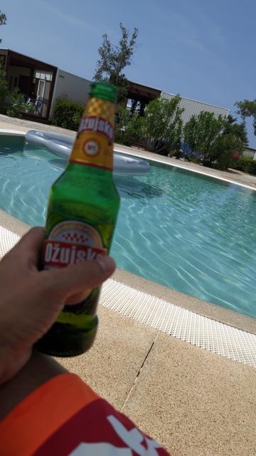 Jemand hält eine Flasche Ožujsko Bier in der Hand, dahinter ein Pool mit türkisblauem Wasser, Oleanderastauden, Mobilheime und blauem Himmel