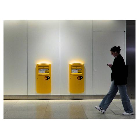 The image depicts an indoor railway station setting with a person walking past two yellow mail boxes mounted on a wall. The individual is dressed in casual attire, including a dark jacket, light blue jeans, and white sneakers, and appears to be looking at a smartphone. The background consists of a plain, light-colored wall and a tiled floor. (created with help of Mistral.ai)