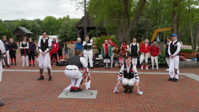 Four morris dancers (plus the arm and foot of a fifth) with white shirts and trousers and black vests, decorated with multicolor ribbons, dancing on a brick paved surface. Two are standing and two are crouched low. Behind them an audience including some other morris dancers.