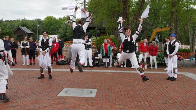 Four morris dancers (plus the arm and leg of a fifth) with white shirts and trousers and black vests, decorated with multicolor ribbons, dancing on a brick paved surface. Two are standing and two are leaping high, arms raised. Behind them an audience including some other morris dancers.