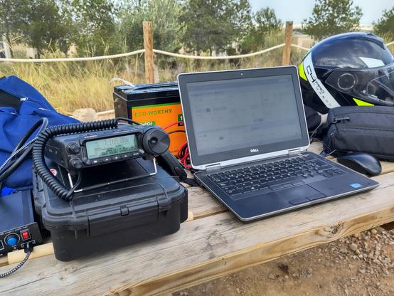 Wooden table with my Amateur Radio equipment. From left to right: A Ham Radio, a Yaesu FT-891 on top of a carrying case, at his side, an Antenna Tuner. Behind, a car-sized battery. To the right a Laptop, and then a Motorcycle helmet. In the background some rope between poles make a fence to a field and a couple of trees.