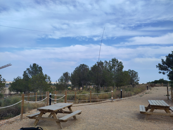 Pictured a park. there is a path with a couple of picnic tables, ant the sides of the path a rope fence with posts, separating unmantained grass fields and trees. At the center of the picture a fishing pole can be seen, sustaining a wire that forms an inverted vee shape.