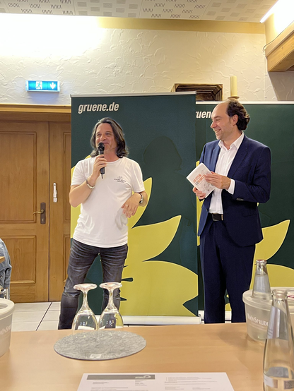 Das Bild zeigt zwei Männer in einem Raum, der offensichtlich für eine Veranstaltung oder eine Pressekonferenz eingerichtet ist. Der Mann links trägt ein weißes T-Shirt und Jeans und hält ein Mikrofon in der Hand, während er spricht. Der Mann rechts trägt einen dunkelblauen Anzug und hält ein Papier in der Hand. Hinter ihnen ist ein Banner mit der Aufschrift "gruene.de" zu sehen, was auf die Partei Die Grünen hinweist. Das Banner zeigt auch eine gelbe Blume, die ein Symbol der Partei ist. Vor ihnen steht ein Tisch mit zwei umgekehrten Weingläsern und einer Wasserflasche. Die Atmosphäre wirkt informell und freundlich.