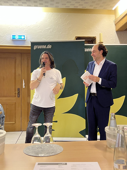 Das Bild zeigt zwei Männer in einem Raum, der offensichtlich für eine Veranstaltung oder eine Pressekonferenz eingerichtet ist. Der Mann links trägt ein weißes T-Shirt und Jeans und hält ein Mikrofon in der Hand, während er spricht. Der Mann rechts trägt einen dunkelblauen Anzug und hält ein Papier in der Hand. Hinter ihnen ist ein Banner mit der Aufschrift "gruene.de" zu sehen, was auf die Partei Die Grünen hinweist. Das Banner zeigt auch eine gelbe Blume, die ein Symbol der Partei ist. Vor ihnen steht ein Tisch mit zwei umgekehrten Weingläsern und einer Wasserflasche. Die Atmosphäre wirkt informell und freundlich.