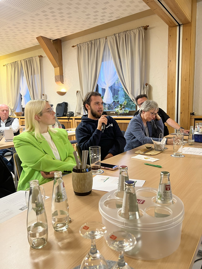 Das Bild zeigt eine Gruppe von Personen in einem gemütlichen, möglicherweise traditionellen Raum, der mit Holzbalken und großen Fenstern mit weißen Vorhängen ausgestattet ist. Die Personen sitzen an einem langen Holztisch, auf dem mehrere Flaschen Wasser, Gläser und Notizblätter verteilt sind. Ein Mann in einem dunklen Sweatshirt hält ein Mikrofon und scheint zu sprechen, während eine Frau in einem leuchtend grünen Blazer neben ihm aufmerksam zuhört. Eine weitere Frau in einem blauen Blazer sitzt ebenfalls am Tisch und schaut nachdenklich in die Ferne. Im Hintergrund sind weitere Personen zu sehen, die ebenfalls an dem Gespräch teilnehmen. Die Atmosphäre wirkt konzentriert und professionell, was auf eine formelle Besprechung oder ein Treffen hinweist.