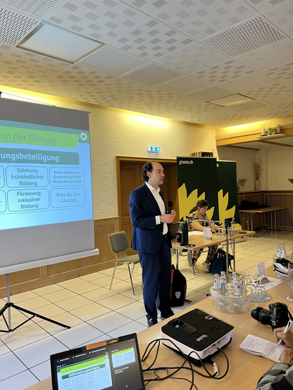 Das Bild zeigt eine Präsentation in einem Konferenzraum. Ein Mann in einem dunkelblauen Anzug hält eine Rede und hält ein Mikrofon in der Hand. Er steht vor einer Projektionsfläche, auf der ein PowerPoint-Präsentationsfolie mit dem Titel "Bildung" und dem Untertitel "Bildungsbeteiligung" zu sehen ist. Die Folie enthält verschiedene Punkte wie "Stärkung frühkindlicher Bildung", "Förderung inklusiver Bildung", "beste Schulen an Orten mit größten Herausforderungen" und "Pakt für den Ganztag". Im Hintergrund sitzt eine Frau an einem Tisch, der mit Wasserflaschen, Gläsern und einem Laptop ausgestattet ist. Auf dem Tisch steht auch ein Projektor. Die Wände des Raumes sind mit hellen Farben gestrichen, und der Boden ist mit weißen Fliesen ausgelegt. Ein Banner mit dem Text "gruene.de" ist sichtbar.