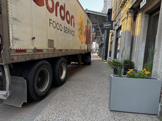 An 18-wheel truck is parked up on the sidewalk leaving little room while delivering boxes of crappy food to restaurants