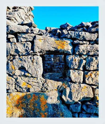 This photo shows a section of a weathered stone wall against a bright blue sky.  The wall is made of rough, irregularly shaped stones, many showing signs of age and discoloration.  Patches of orange lichen are visible on some of the stones.  A small, dark opening, a window, is visible in the center of the wall.