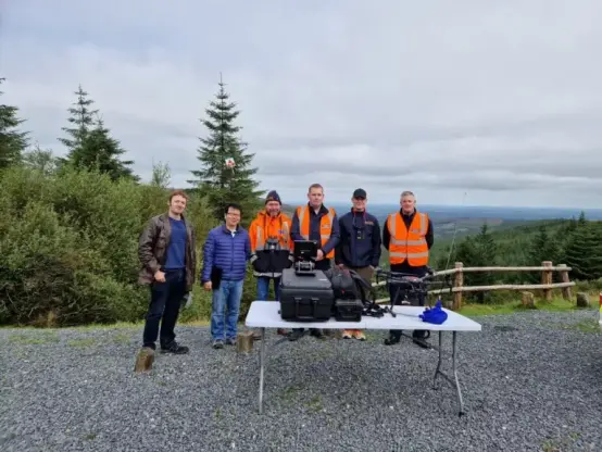 A group of 6 men. Outdoors in the mountains with trees, on a table radio and drones
