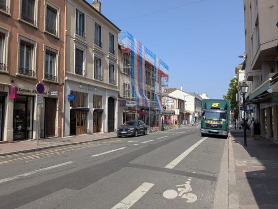 Bestandsstraßenraum der Avenue des frères Lumière zwischen Rue Saint-Gervais und Rue Villon, wo der Umbau noch nicht begonnen hat. Es gibt auf beiden Seiten gerade so 2 Meter breite Bürgersteige, in der Mitte eine etwa 9 m breite Fahrbahn mit markierten Radstreifen. Ein LKW parkt auf dem Radstreifen.