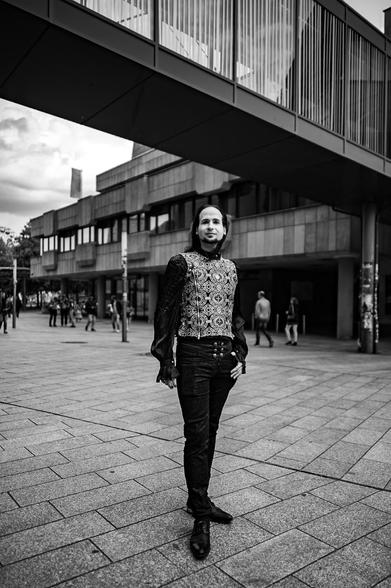 In the stark monochrome, a solitary figure stands on the brick-paved expanse before what appears to be a municipal building in Europe. The figure commands attention, their presence amplified by their attire. Behind them, other humans walk around the streets. Overhead, a modern skybridge casts an imposing shadow, a stark contrast to the scene unfolding below.