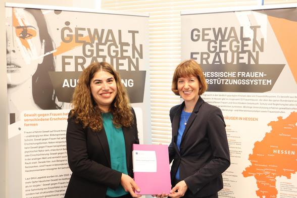 Auf dem Foto sind zwei lächelnde Personen zu sehen, die eine gedruckte Publikation in die Kamera halten. Im Hintergrund sind Aufsteller mit der Überschrift "Gewalt gegen Frauen".