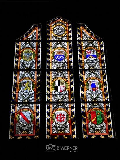 Kirchenfenster in der Schlosskirche Wittenberg mit farbiger Glasmalerei und Wappen verschiedener Städte, darunter Altdorf, Erlangen, Kulmbach und Mühlhausen. Das Fenster ist reich verziert mit floralen Mustern und goldenen Rahmen, die die kunstvollen Stadtwappen hervorheben.