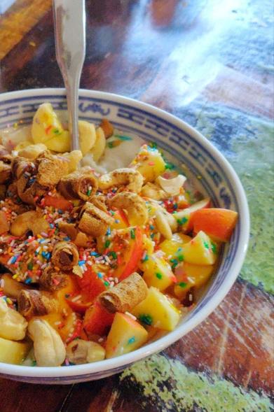 Dunkler Tisch, blaueweiße asiatische Porzellanschale, darin Porridge mit allerlei Toppings (Banane, Apfel, Schokokeksröllchen, Cashews, Zuckerstreusel, Zimt), ein Löffel steckt im Brei.