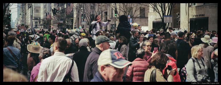 Two people sit on the shoulders of others above a large crowd of people.  One is directing the other with a phone in their hand; both are prepared to take photos of one another.