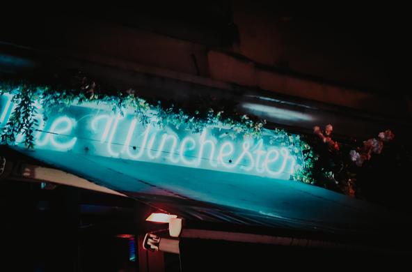 Front néons of the Winshester bar : cold blue, radiating on the store and ivy besides it. Manuscript font. The wall behind is dark warm tones