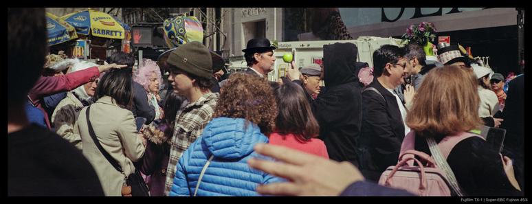 A person wearing a bowler hat and green apple suspended in front of it, as in the painting "The Son of Man", is visible through a throng of people at a parade.