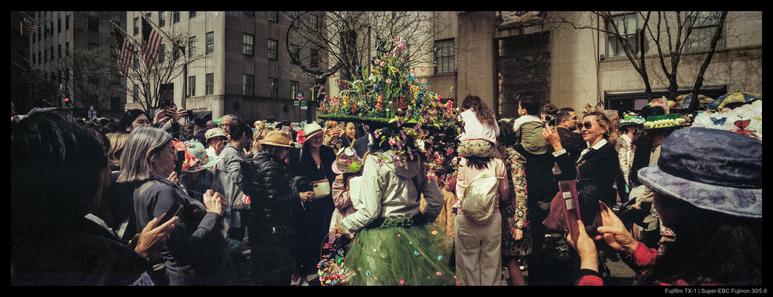 A person in a green dress and an enormous flowery and bedecked hat stands ringed by a crowd of interested people, many of whom have their phones raised to take a picture.