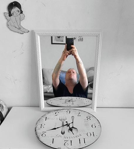 A person is taking a selfie using a mirror placed on top of a white dresser. Their arms are raised high holding a phone, and their reflection shows a focused expression. The mirror is framed in white and reflects part of a couch and wall decor behind them. On the dresser in front of the mirror is a decorative clock with vintage-style utensils as clock hands. The room is mostly monochrome with soft lighting, and a small illustration of a cartoon-style angel is visible on the wall above the mirror.