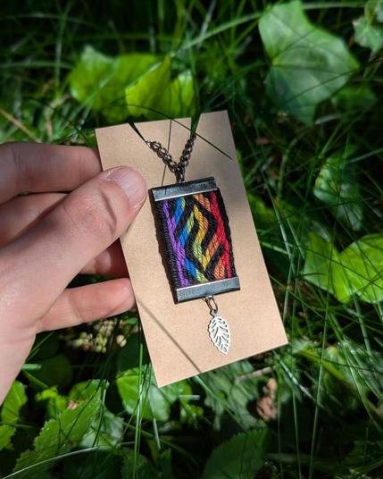 Photo d'un pendentif porte-clés tissé aux couleurs du drapeau arc-en-ciel sur une cartonnette de présentation tenue par une main sur fond d'herbe et de lierre.