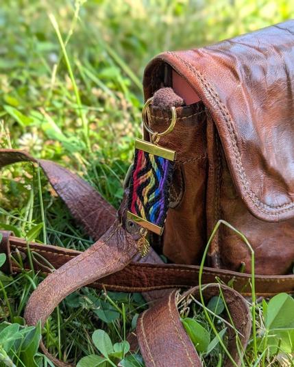 Photo d'un porte-clés tissé aux couleurs du drapeau arc-en-ciel accroché à un sac en cuir posé sur l'herbe.