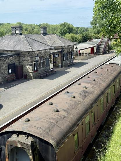 A train carriage seen from above. On tracks opposite an old Victorian train station. Trees in the distance.
