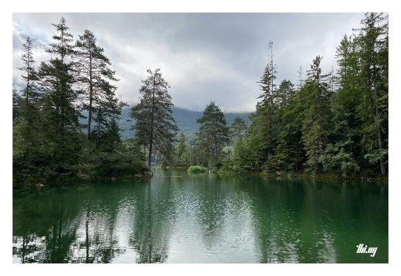 Photo of a small bay of a large mountain lake with rick green water, surrounded by lush mixed alpine forest. A small island (also forested) is further into the lake, outside the bay. A dense layer of gray, moody, deep hanging clouds is obstructing the view of the tall peaks in the background.