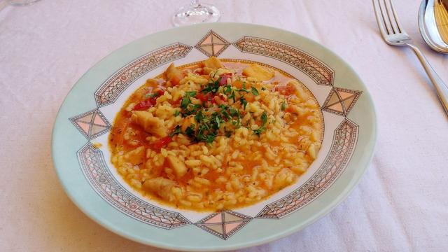 Un plato hondo con arroz caldoso con sepia y calamar adornado con un poco de perejil picado. En la cuchara que hay al lado del plato puede verse reflejado el toldo de la pérgola.