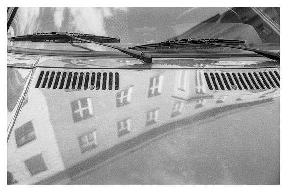 Black and white photograph of a reflection on the hood of an Audi 80. The picture shows part of the windscreen with the windscreen wipers and the steering wheel, as well as part of the hood with the air inlets. A house front with windows is reflected in the hood.