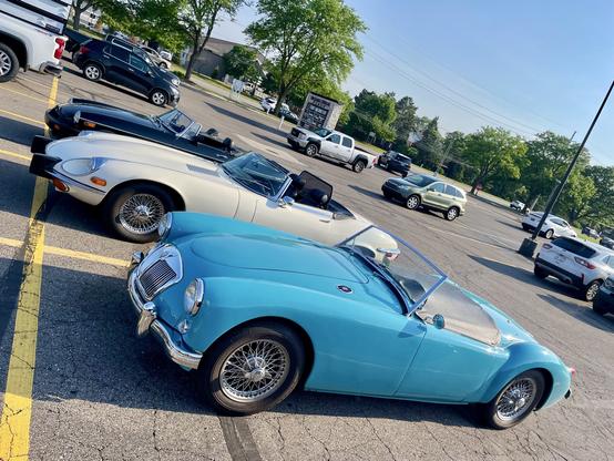 Photograph of three classic British sports cars in a parking lot with modern cars in the background. A blue MGA is in front with a white Jaguar E-Type behind and a black MGB behind that. The photograph tilts to the lower right.
