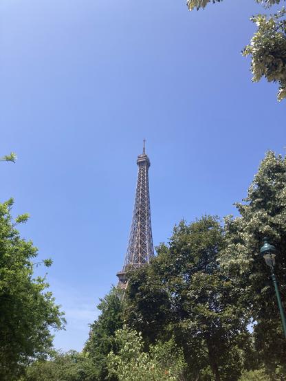 Photo de la tour Eiffel vue à travers les arbres (au premier plan). Il y a un grand ciel bleu