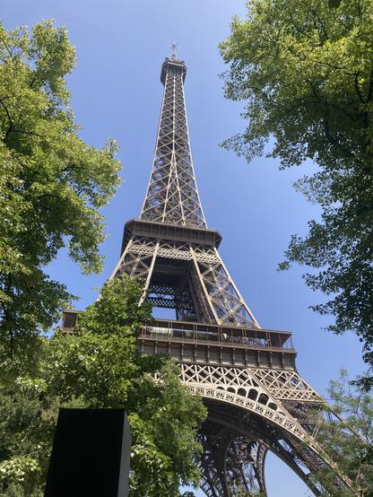 Vue de la tour Eiffel depuis son pied ; à droite et à gauche, des arbres. Le ciel est bleu