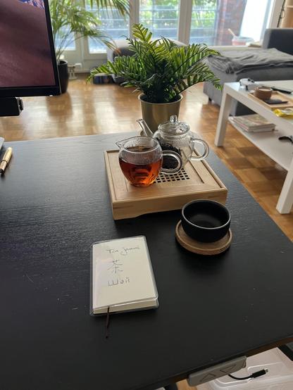 photo of a tea set next to a small journal. the journal has ‘tea’ hand-written in english, japanese, and kazakh on the front. the small glass teapot is filled with a dark red-brown tea