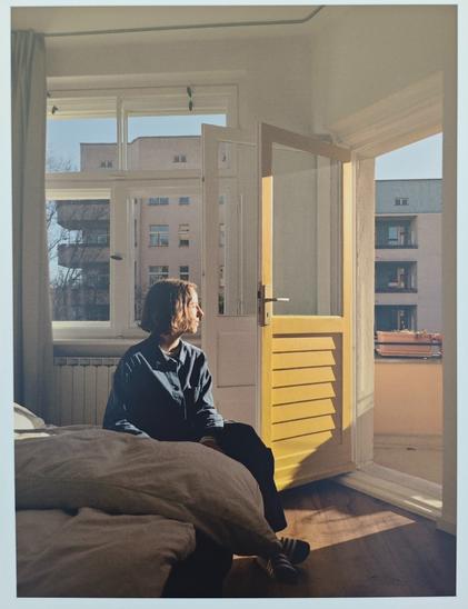 person sitzt im schlafzimmer, wohl in einem hochhaus, es ist auch ein hochhaus gegenüber. die balkontüre ist offen und wird von warmer sonne angeschienen