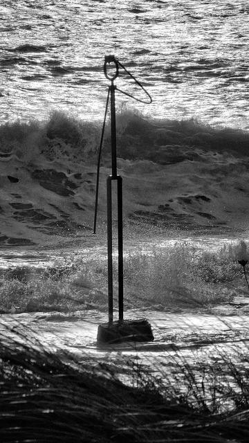 A black and white photograph of a tall, dark, abstract metal sculpture of a man saluting, stands firmly on a rocky base at the edge of a turbulent sea. Large waves crash behind it, their white foam contrasting sharply with the dark water. The sky is bright and reflects off the choppy surface of the ocean, creating a shimmering effect. In the foreground, dark, blurred blades of grass or reeds are visible along the bottom edge of the frame. The overall mood is dramatic and powerful.