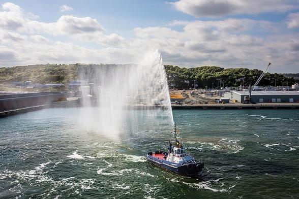 A tugboat sprays water in a display against a backdrop of a coastal industrial area and lush green hills under a partly cloudy sky. The water creates a dramatic plume, and the sunlight enhances the lively scene at the harbor. Photography by Debra Martz