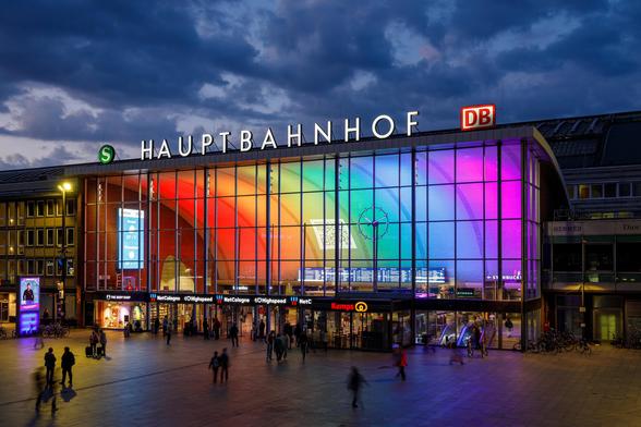 Leicht angeschrägte Dämmerungs-Aufnahme der Empfangshalle des Kölner Hauptbahnhofs von außen. Obendrauf die Buchstaben „Hauptbahnhof“, links davon ein S-Bahn Symbol, rechts das DB-Symbol. Durch die große Fensterfront sieht man in die Empfangshalle, wo die rückwärtige Wand flächendeckend in Regenbogenfarben angestrahlt ist.