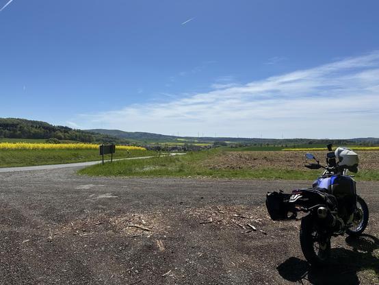A motorcycle parked by a gravel road with a scenic view of rolling hills and a field of yellow flowers under a clear blue sky.