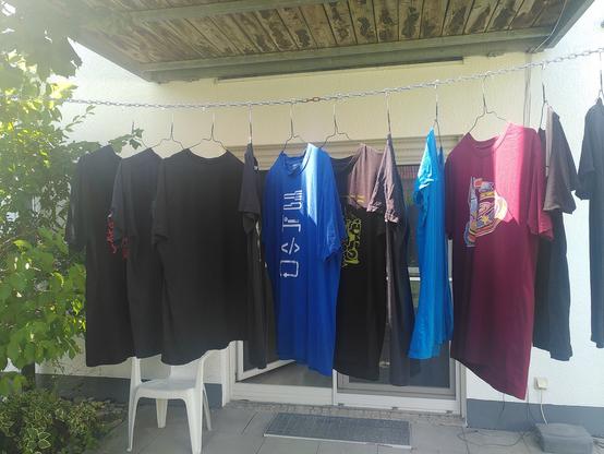 A clothesline stretches between the two supporting pillars of a 1. floor balcony. The Line is a linked metal chain with clothhangers hooked into ever 5th chain link. On the hangers t-shirts of various chaos events are hung for drying. It's dry and sunny day with little wind.