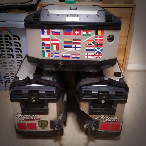 3 metal panniers are stacked in a room. They all have some stickers like flags and the logo of the Africa Twin on it.