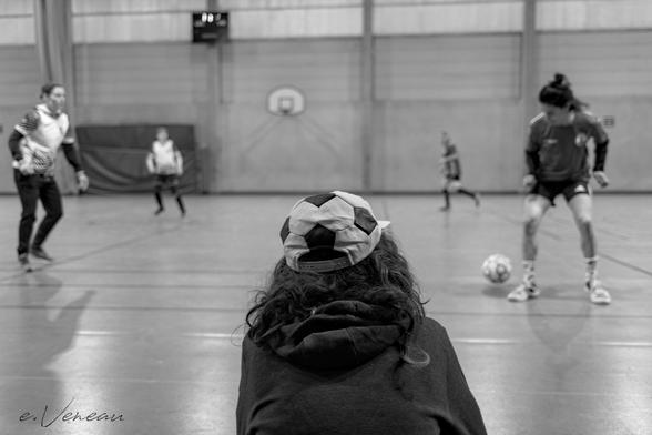 In a gymnasium, four women footballers train on the pitch. On the sidelines, from behind, a woman is watching them: she has a cap on her head in the shape of a football.