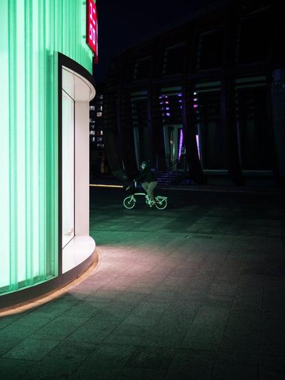 A man on a tiny bicycle cycles past a shop illuminated by green neon light. The rest of the scene is dark.