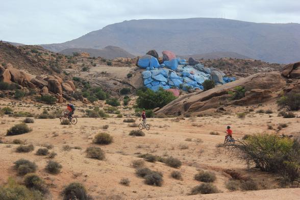 A picture of three people on bicycles, an adult followed in file by two small children, set in a desert landscape. There are scrubby plants sparsely covering the ground. In the distance is a large, flat hill and in the midground a slope of large rocks painted mainly bright blue. These are the Painted Rocks of Tafraoute.
