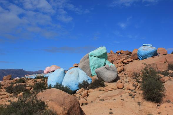 A picture of some of the painted rocks from closer up. there is a mixture of pink, blue and greeny-blue boulders scattered across a shallow slope.