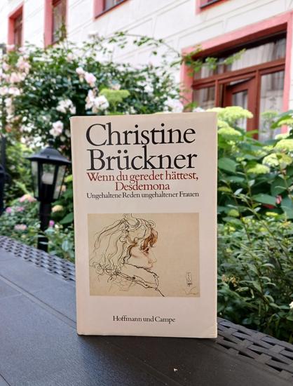 Foto des Buches, auf dessen Cover die bearbeitete Zeichnung »Viola« von Horst Janssen zu sehen ist – eine Frau im Profil mit hochgestelltem Kragen und offenem Haar. Das Buch, dem sein antiquarisches Dasein anzusehen ist, steht auf einer Tischkante vor einem unter anderem mit weißen Rosen und Hortensien bepflanzten Beet vor einer hellen Fassade mit rot umrandeten Fenstern.